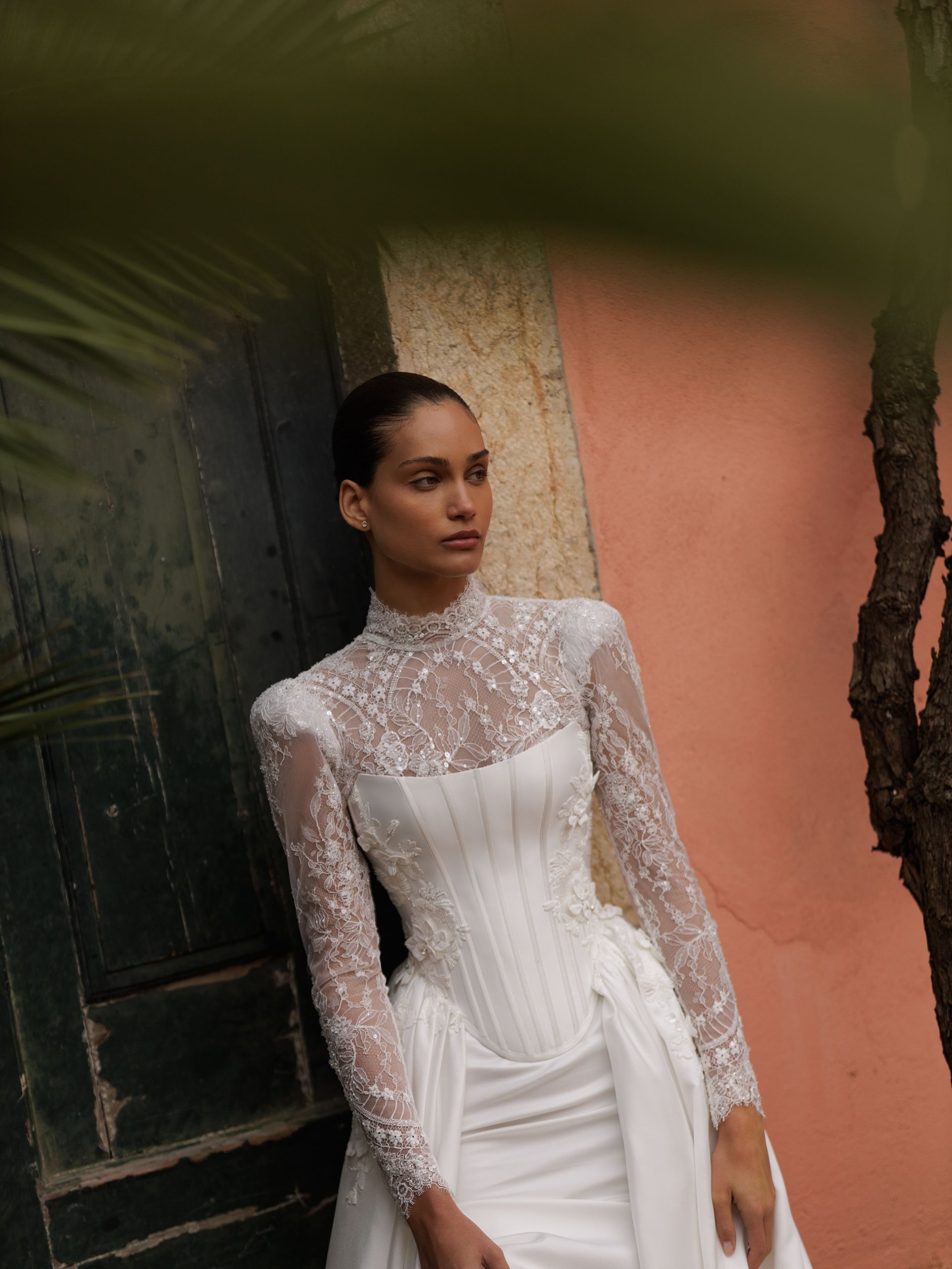 Bride and groom pose on a balcony, holding hands. The bride wears a lace dress and veil, holding a bouquet.