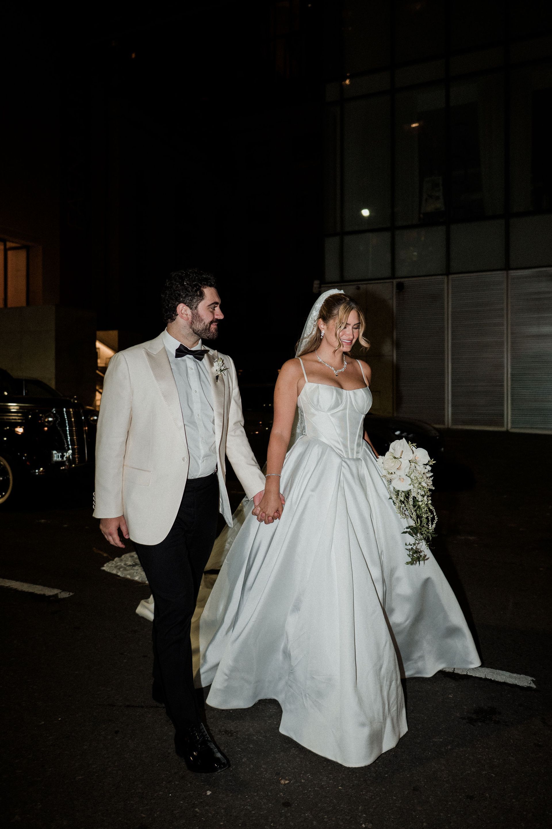 Newlyweds holding hands, exiting a building at night. Bride in satin gown, groom in tuxedo smiling.