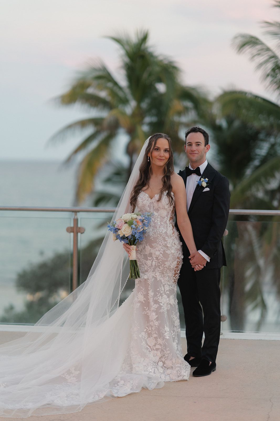 Bride and groom pose on a balcony, holding hands. The bride wears a lace dress and veil, holding a bouquet.