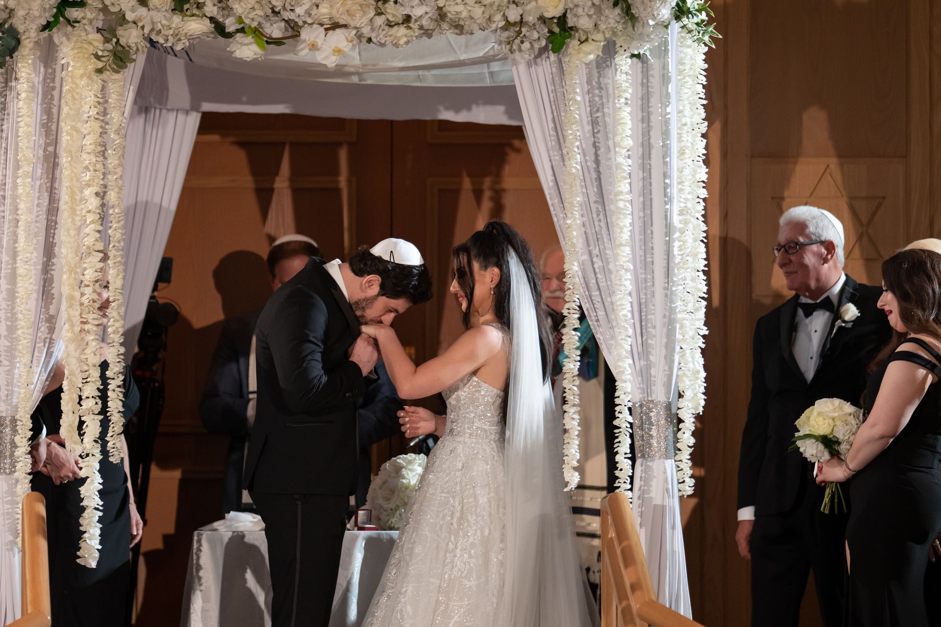 Groom kisses bride's hand under a decorated chuppah at a wedding. Formal attire, white flowers, and guests are present.