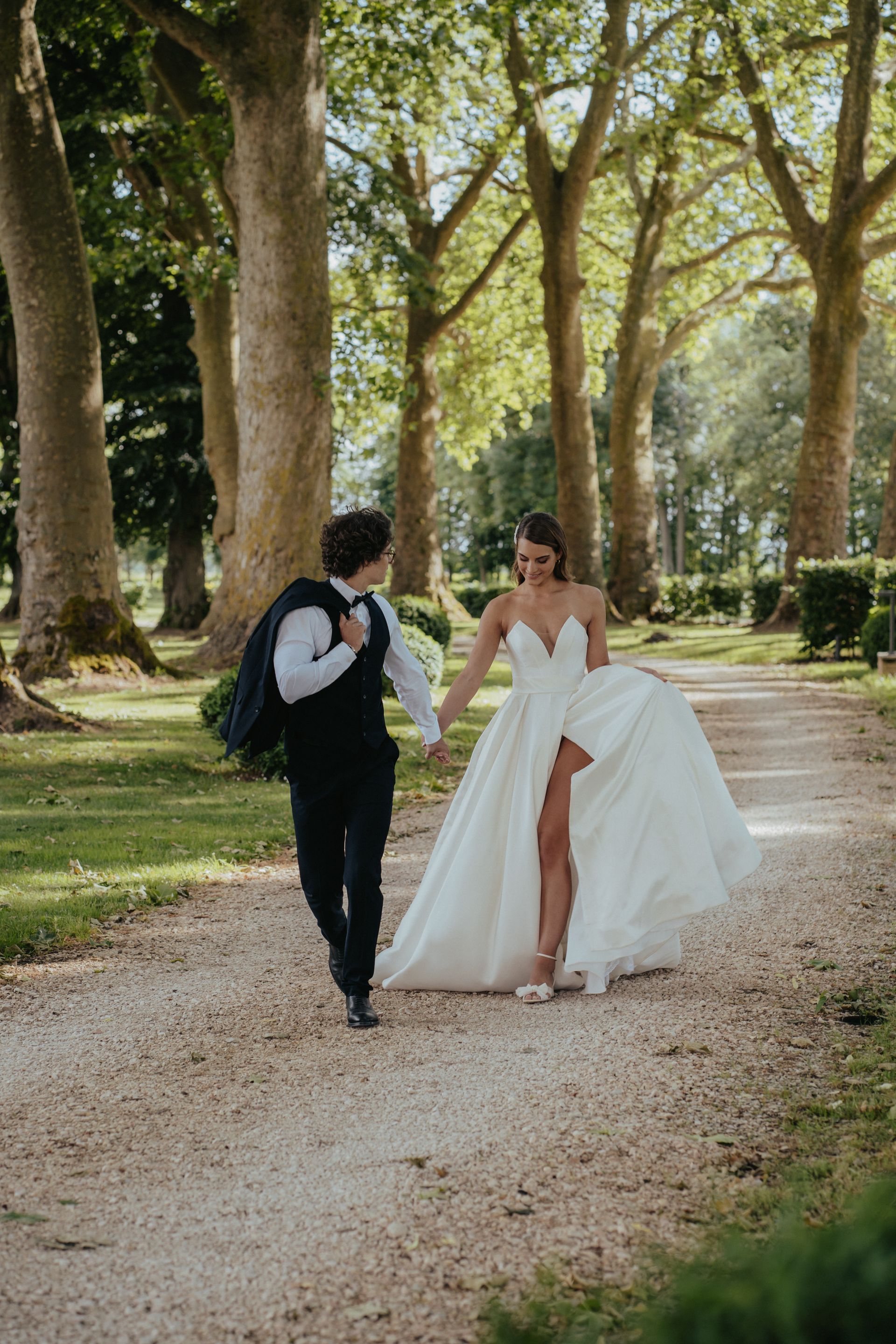 Couple walking hand-in-hand on a path, trees in the background. Bride in a white gown with a slit, groom in a vest.