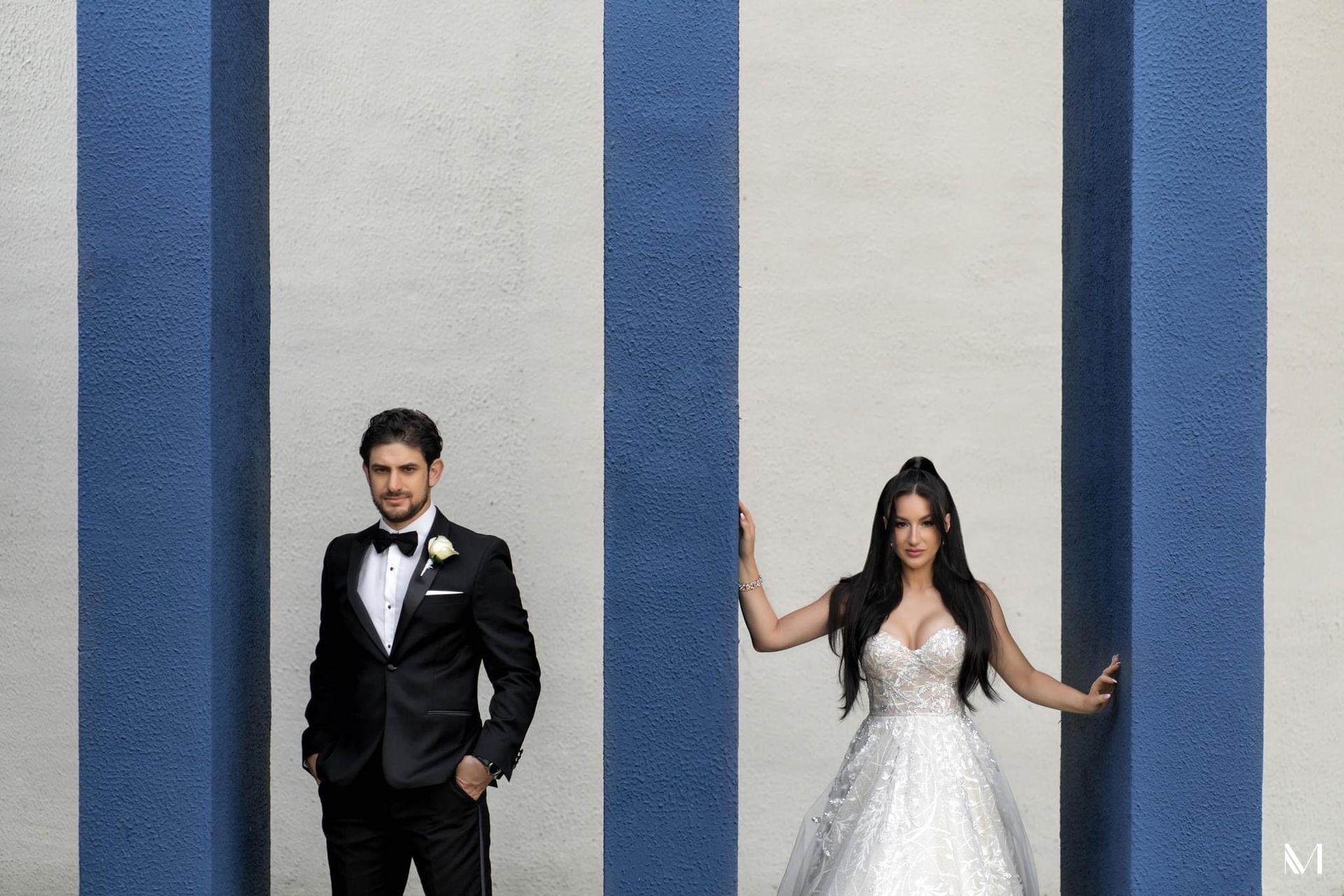 Groom in tux, bride in gown pose between blue pillars. Bride's arms up, groom looks away.