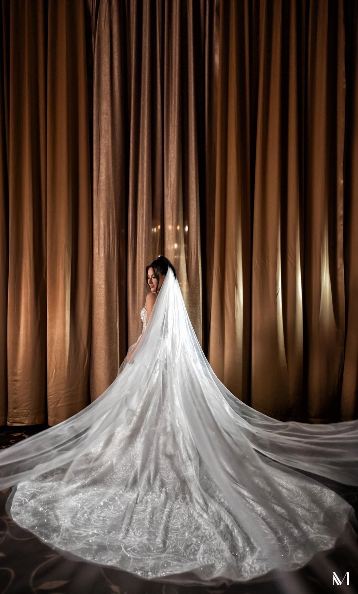 Bride in wedding dress, veil trailing, in front of brown curtains.