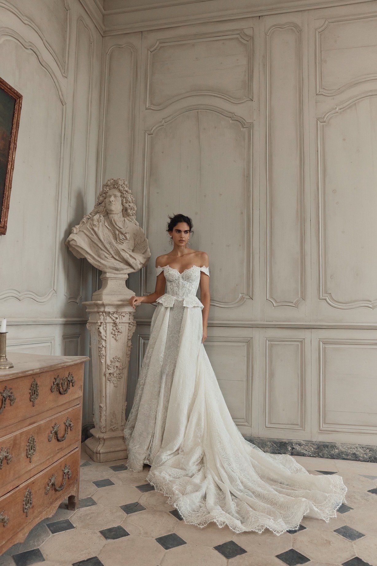 Bride and groom pose on a balcony, holding hands. The bride wears a lace dress and veil, holding a bouquet.