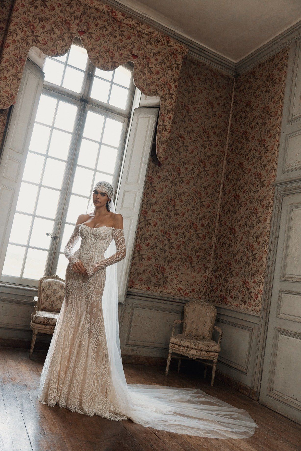 Bride in wedding dress, veil trailing, in front of brown curtains.