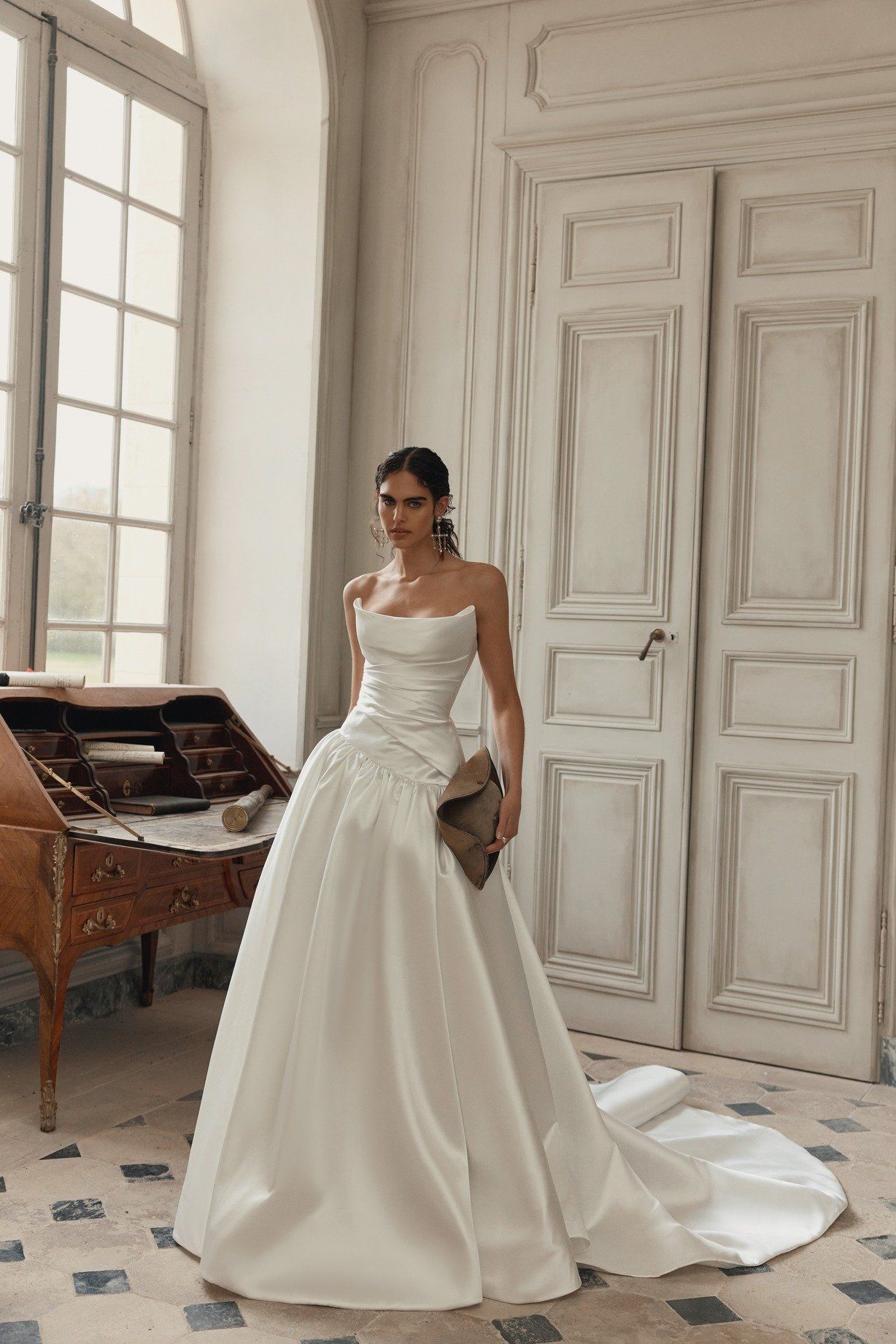 Couple walking hand-in-hand on a path, trees in the background. Bride in a white gown with a slit, groom in a vest.