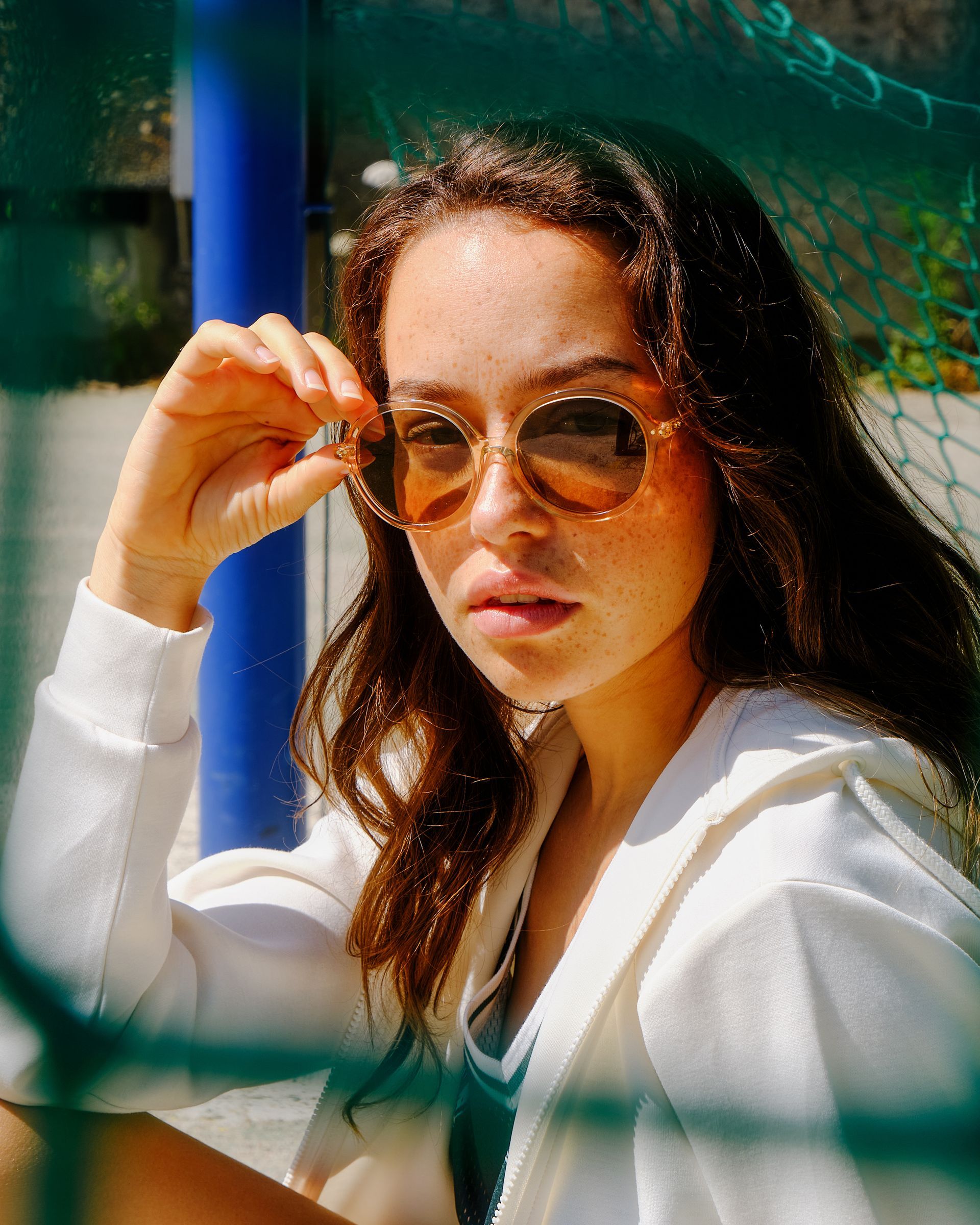 Femme portant des lunettes de soleil et une chemise blanche, appuyée contre un poteau bleu derrière un filet vert.