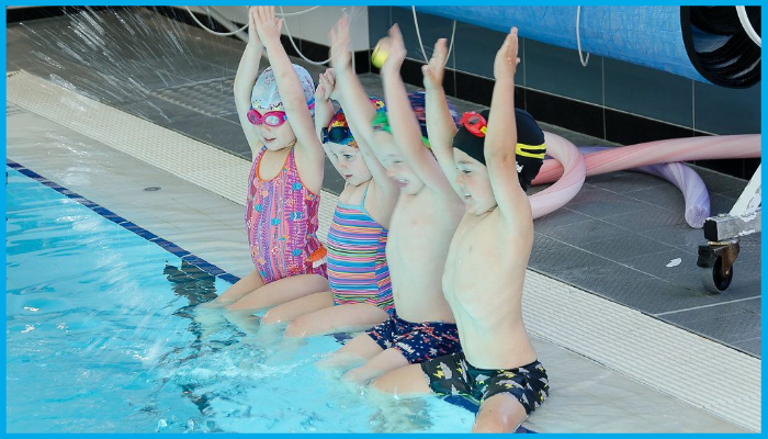 Group of young competitive swimmers sitting on pool edge raising arms in warm-up before training