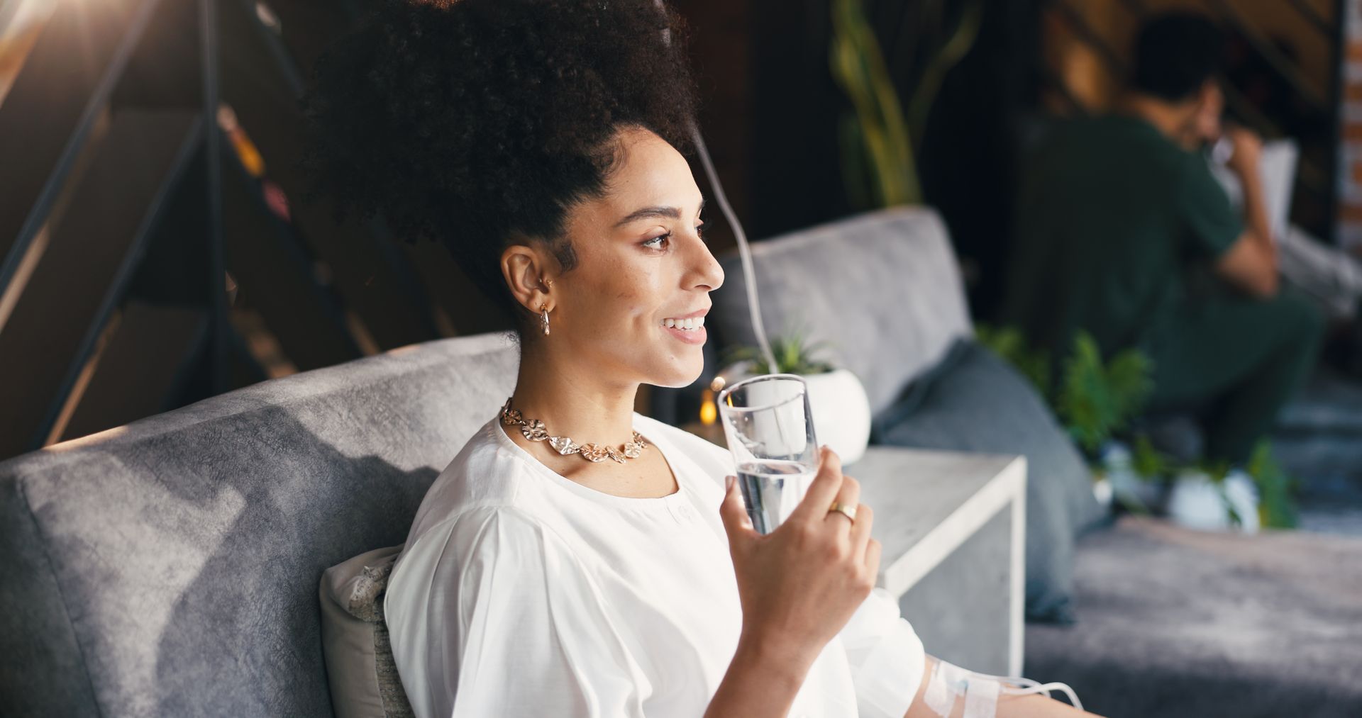 Woman seated on a gray sofa, smiling and holding a glass of water in a bright lounge with an IV in for IV Hydration