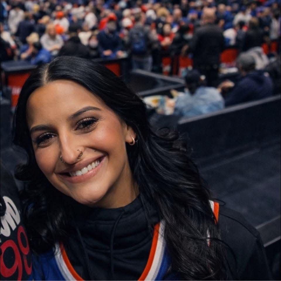 A smiling person with dark hair and a nose ring at a crowded sports arena, wearing a blue and orange jersey. A smiling person with dark hair and a nose ring at a crowded sports arena, wearing a blue and orange jersey.