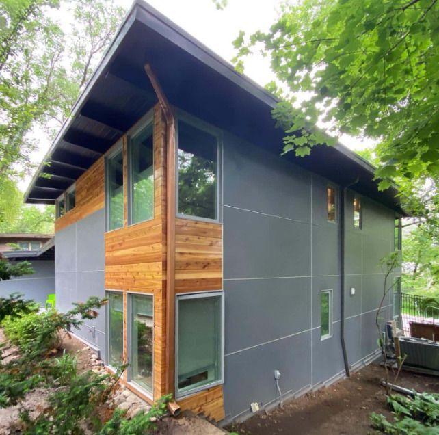 Modern two-story house with gray siding, cedar accents, and large windows set against lush greenery.