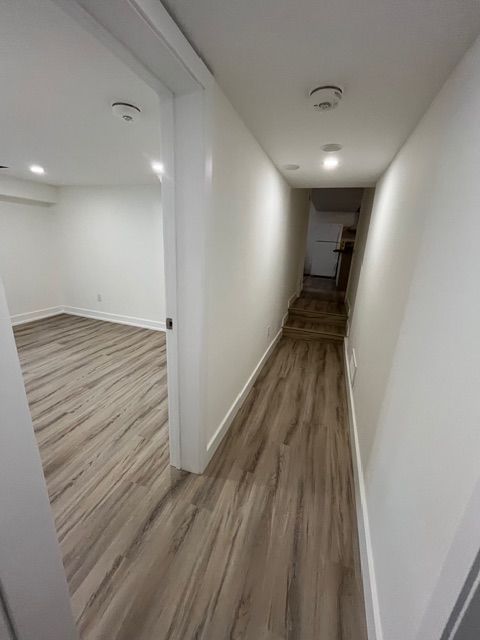 Narrow hallway with white walls and wood-look flooring, leading to stairs. Room to the left has matching flooring.
