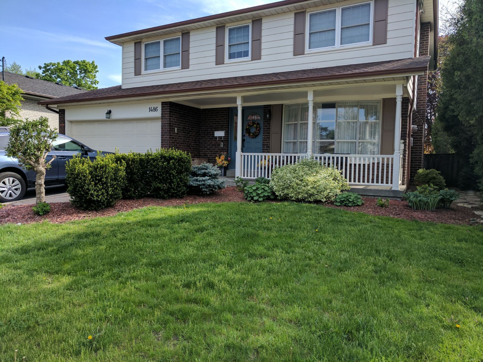 Two-story beige house with white porch and shutters, green lawn, and dark mulch landscaping.