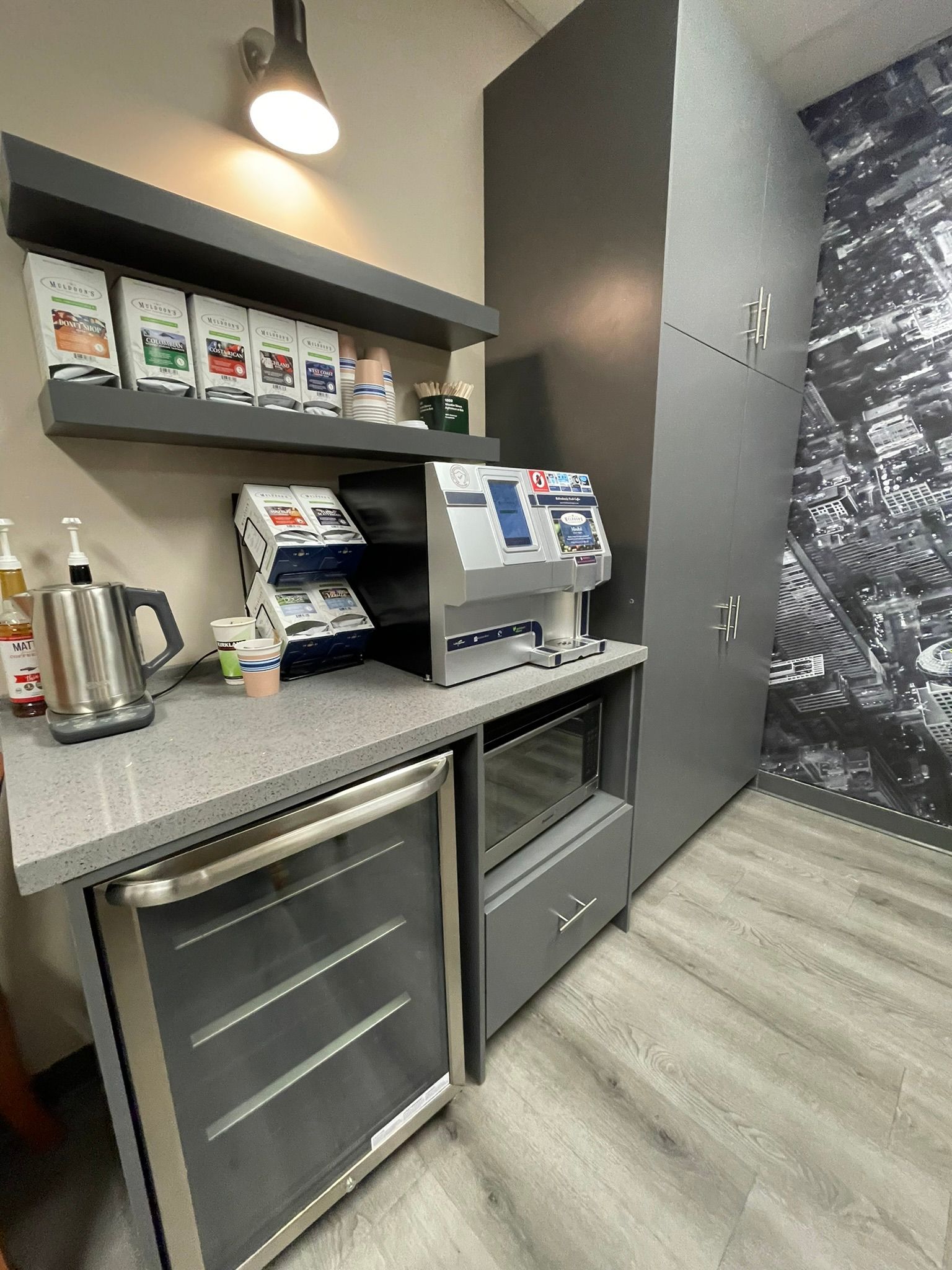Coffee station with grey cabinets, countertop, refrigerator, microwave, and beverage machine.