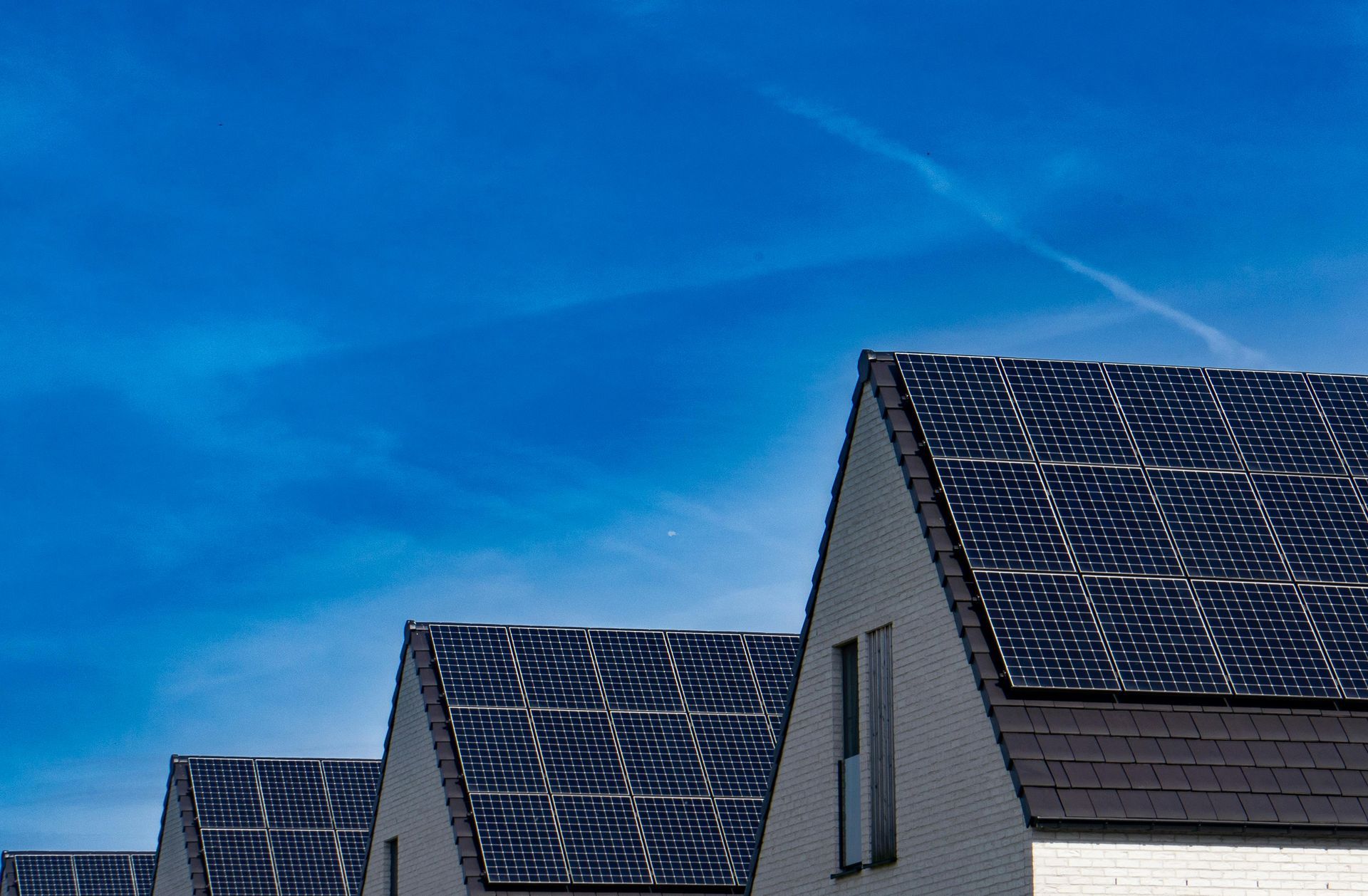 Row of houses with solar panels on rooftops, against a bright blue sky with a few wispy clouds.