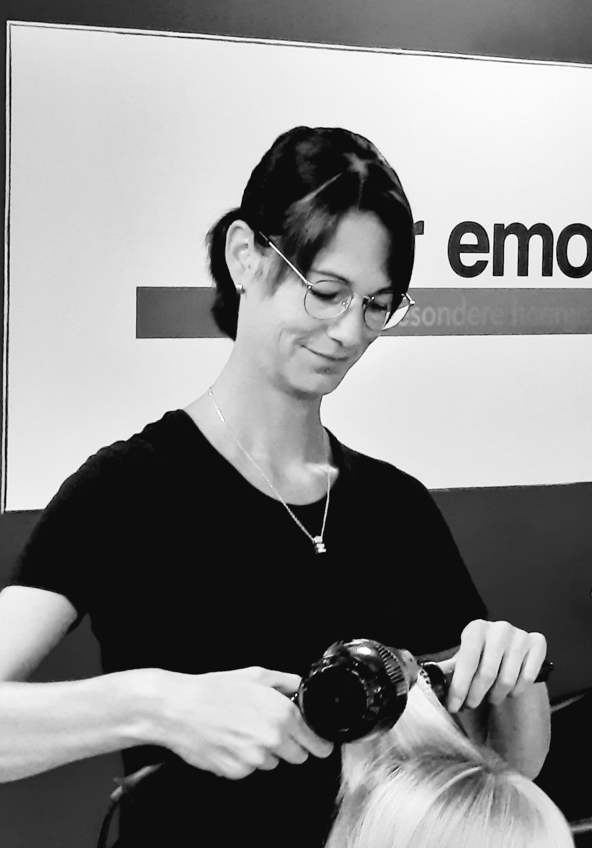 Eine Frau föhnt einer Frau die Haare vor einem Schild mit der Aufschrift „Emo“
