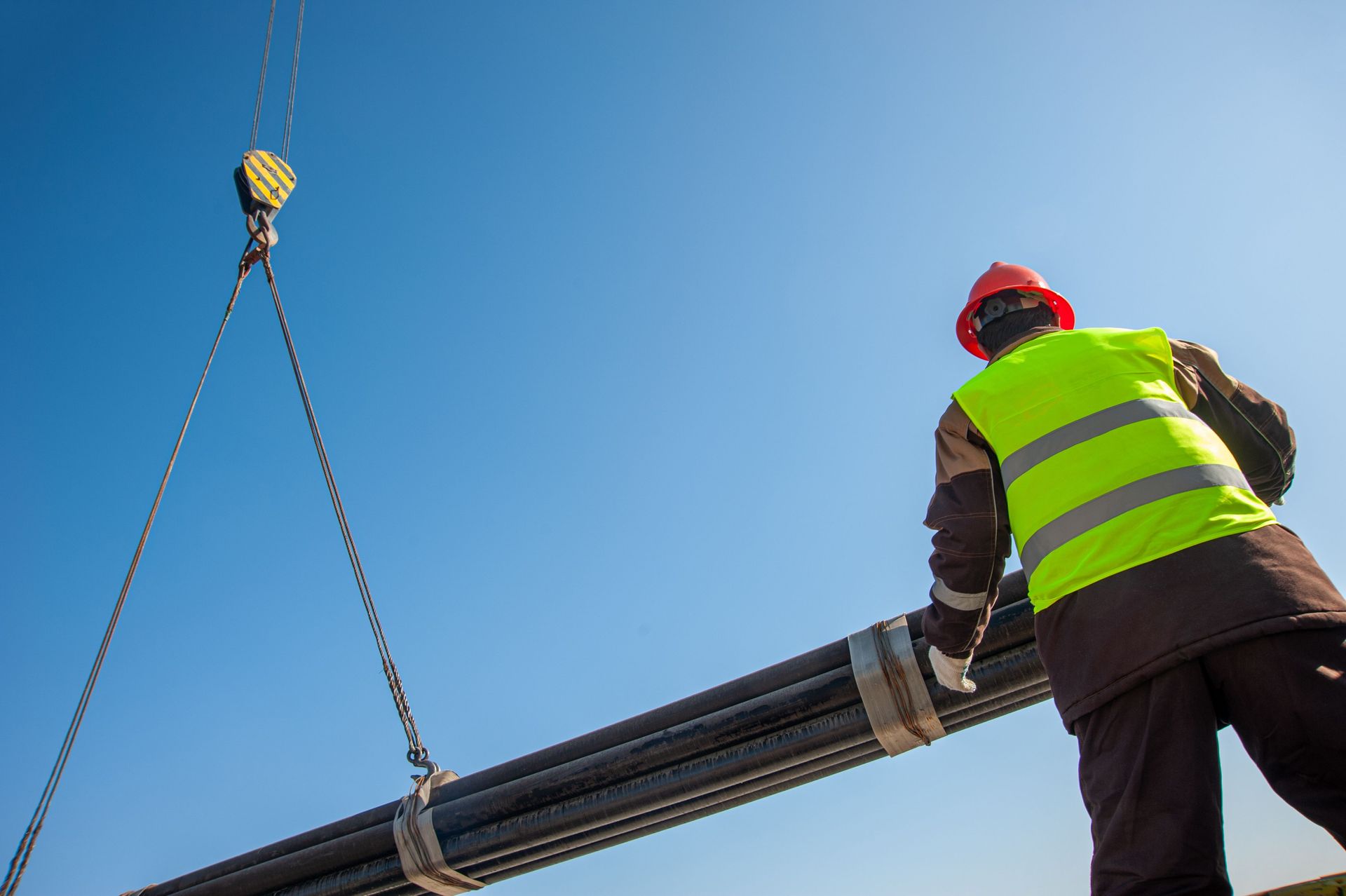 Construction worker in neon vest guiding a crane-lifted bundle of pipes against a blue sky