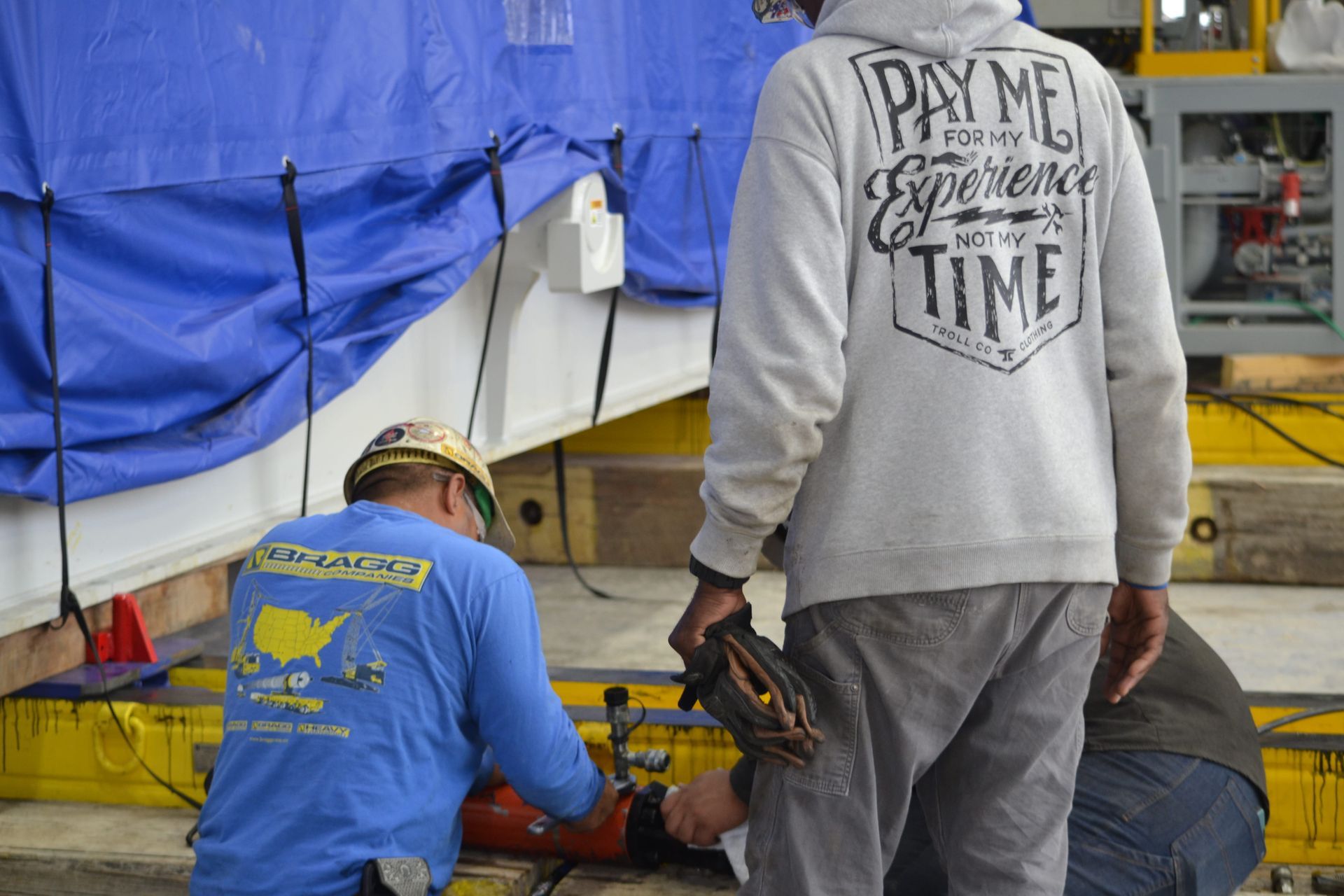 Workers crouch under a blue tarp, using tools beside a gray-shirted person standing nearby.