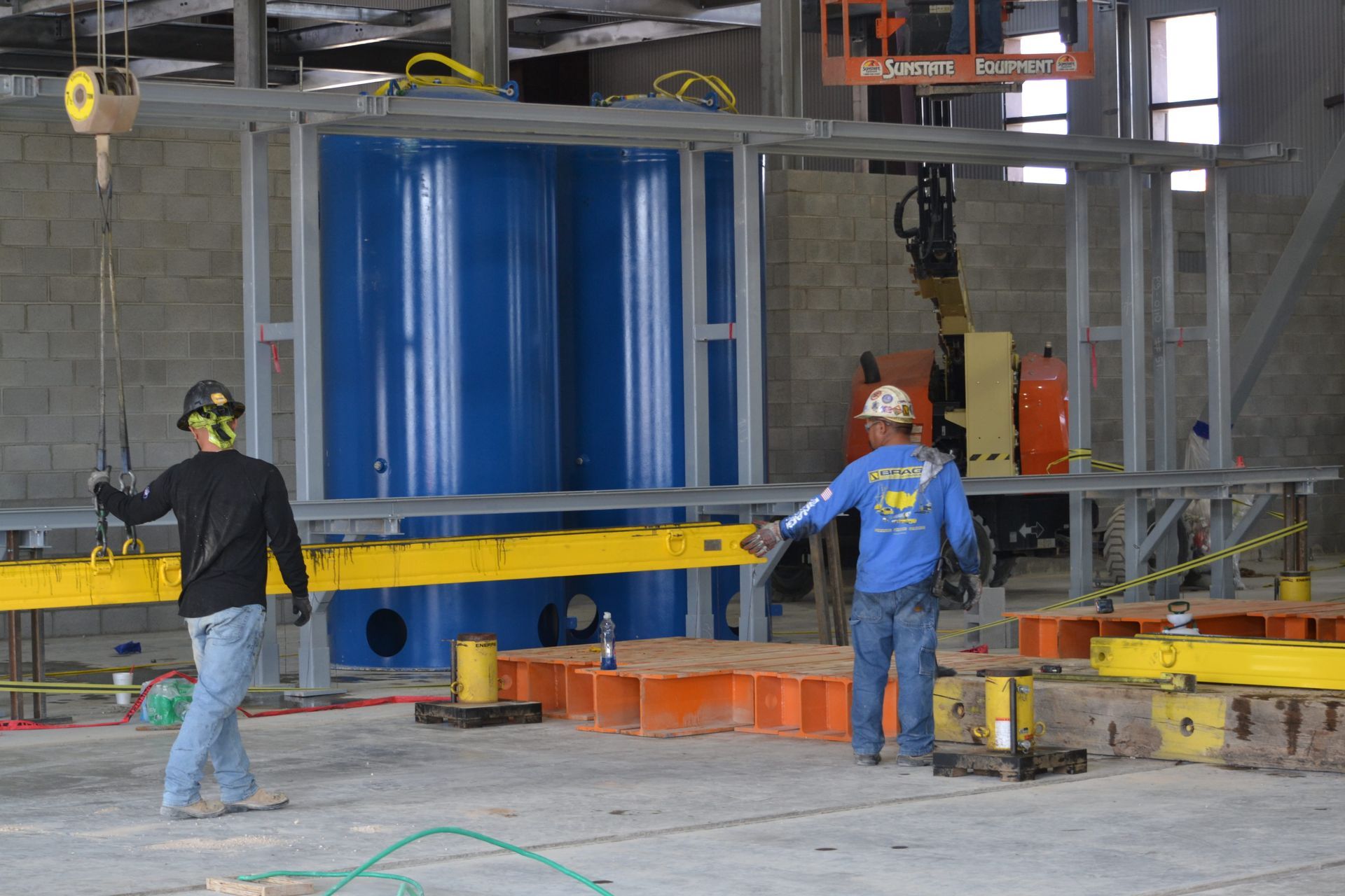 Workers guiding a long yellow beam in an industrial construction site with scaffolding and machinery