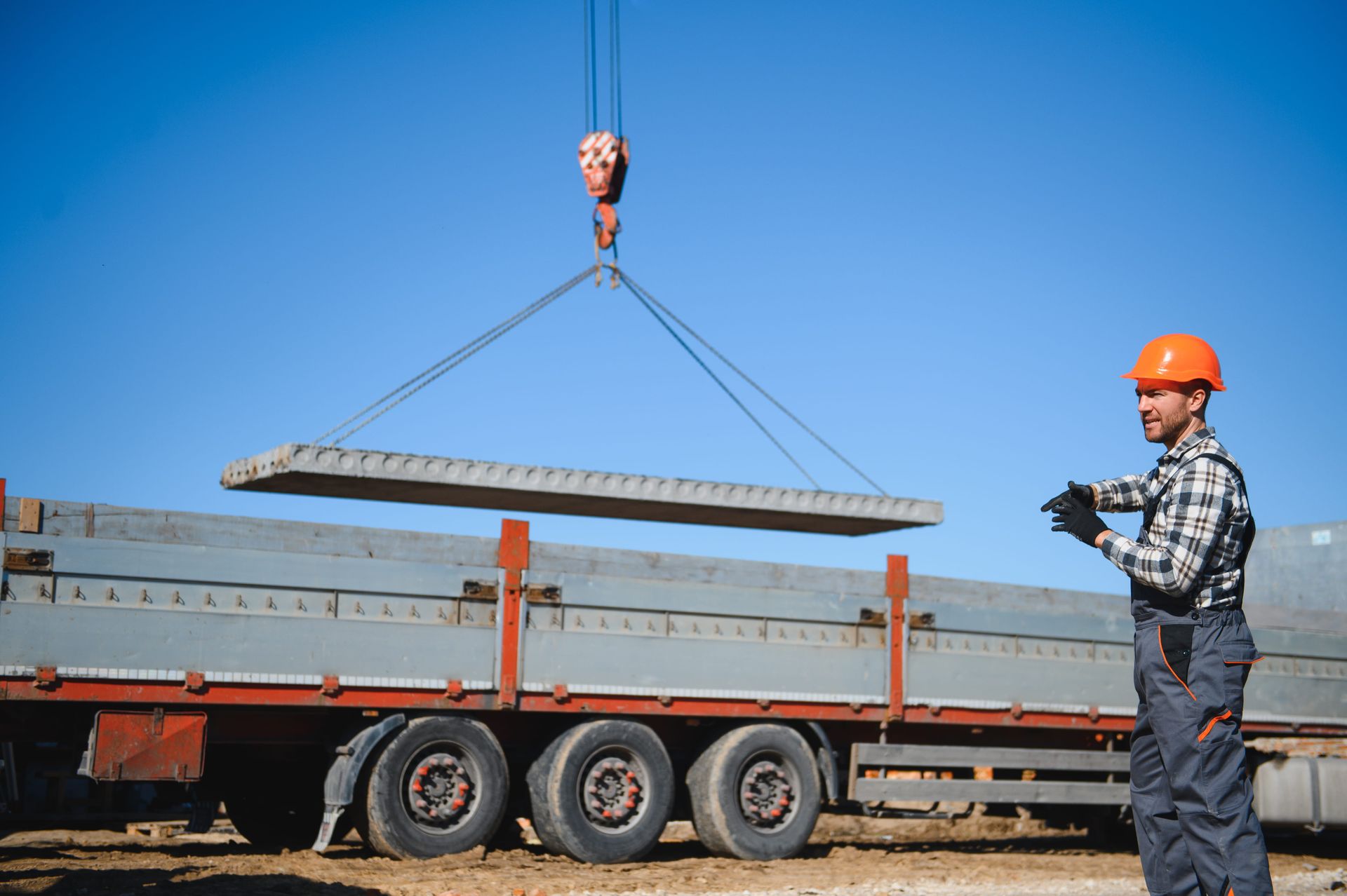 Crane lifting a long steel beam over a flatbed truck while a worker in an orange hard hat guides it.