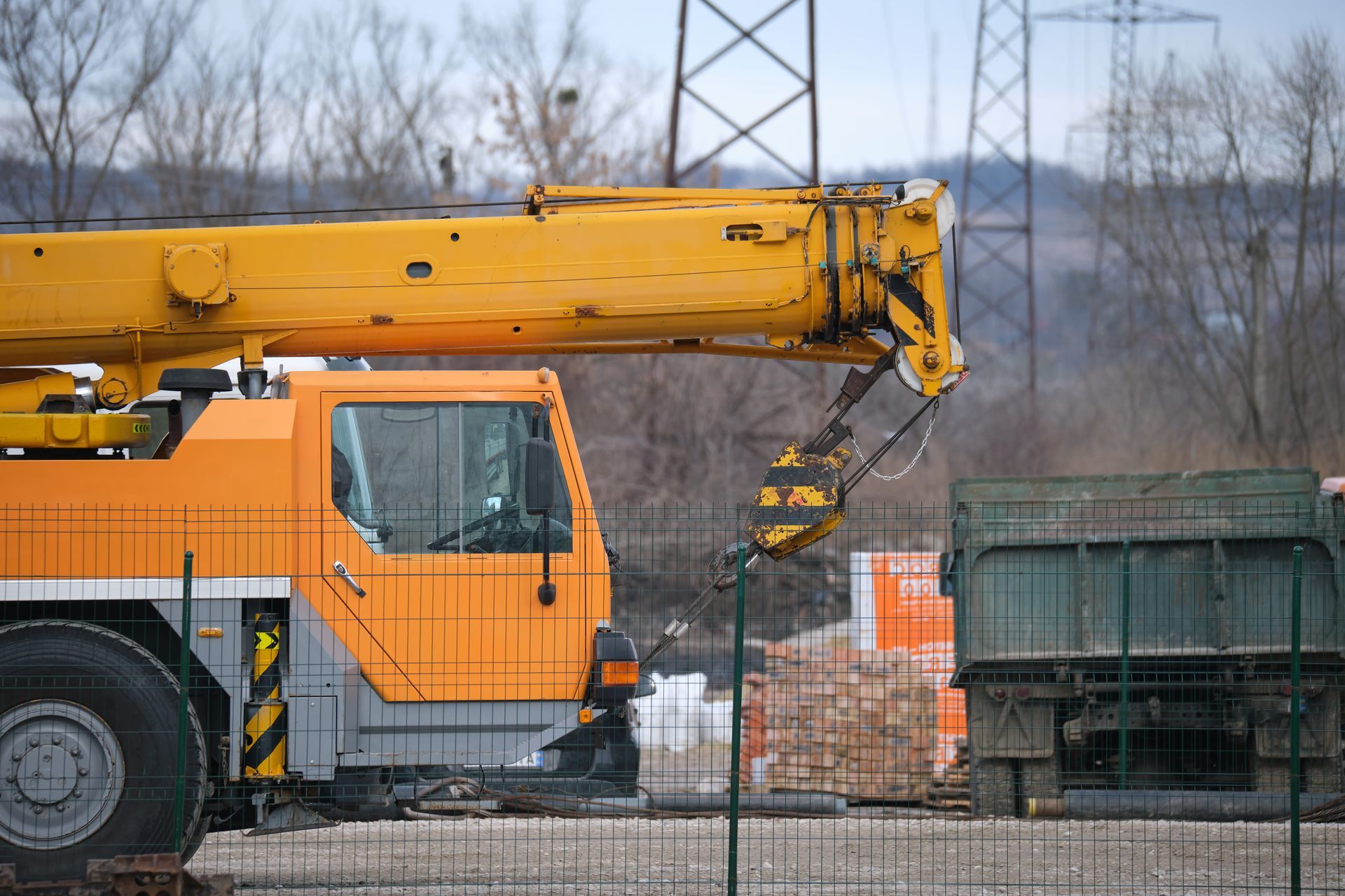 Yellow crane loading debris beside an orange truck near a green dumpster in a fenced industrial area