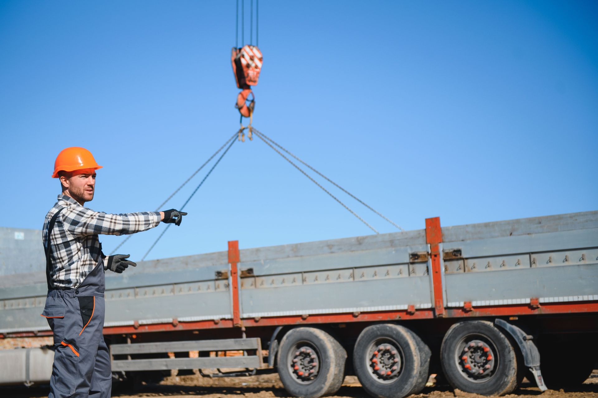Worker in orange hard hat guiding a crane load onto a flatbed trailer under a blue sky