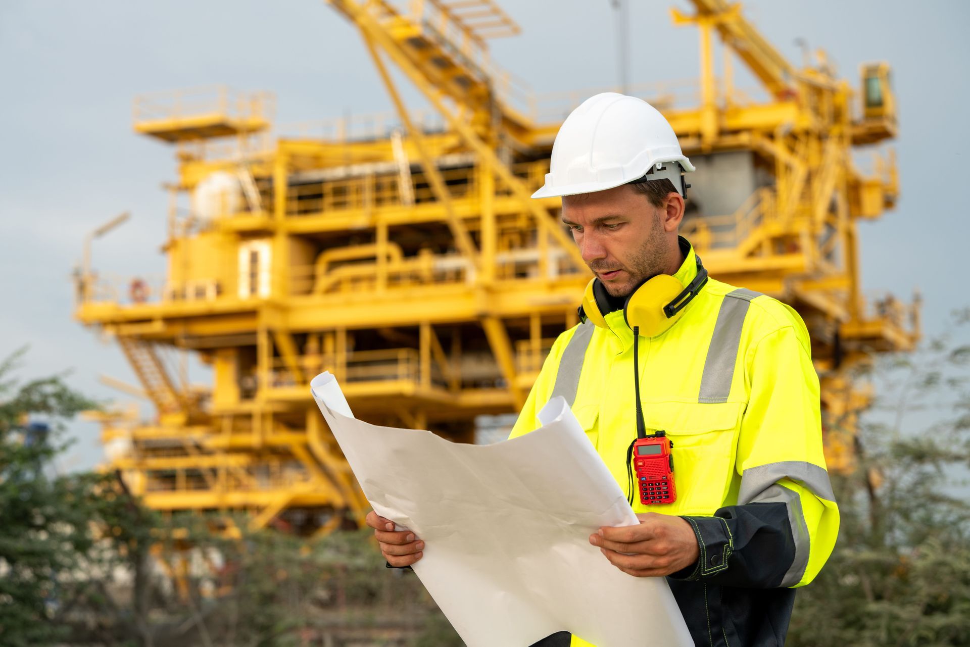 Worker in a white hard hat and neon jacket reviewing plans at an industrial site with yellow cranes.