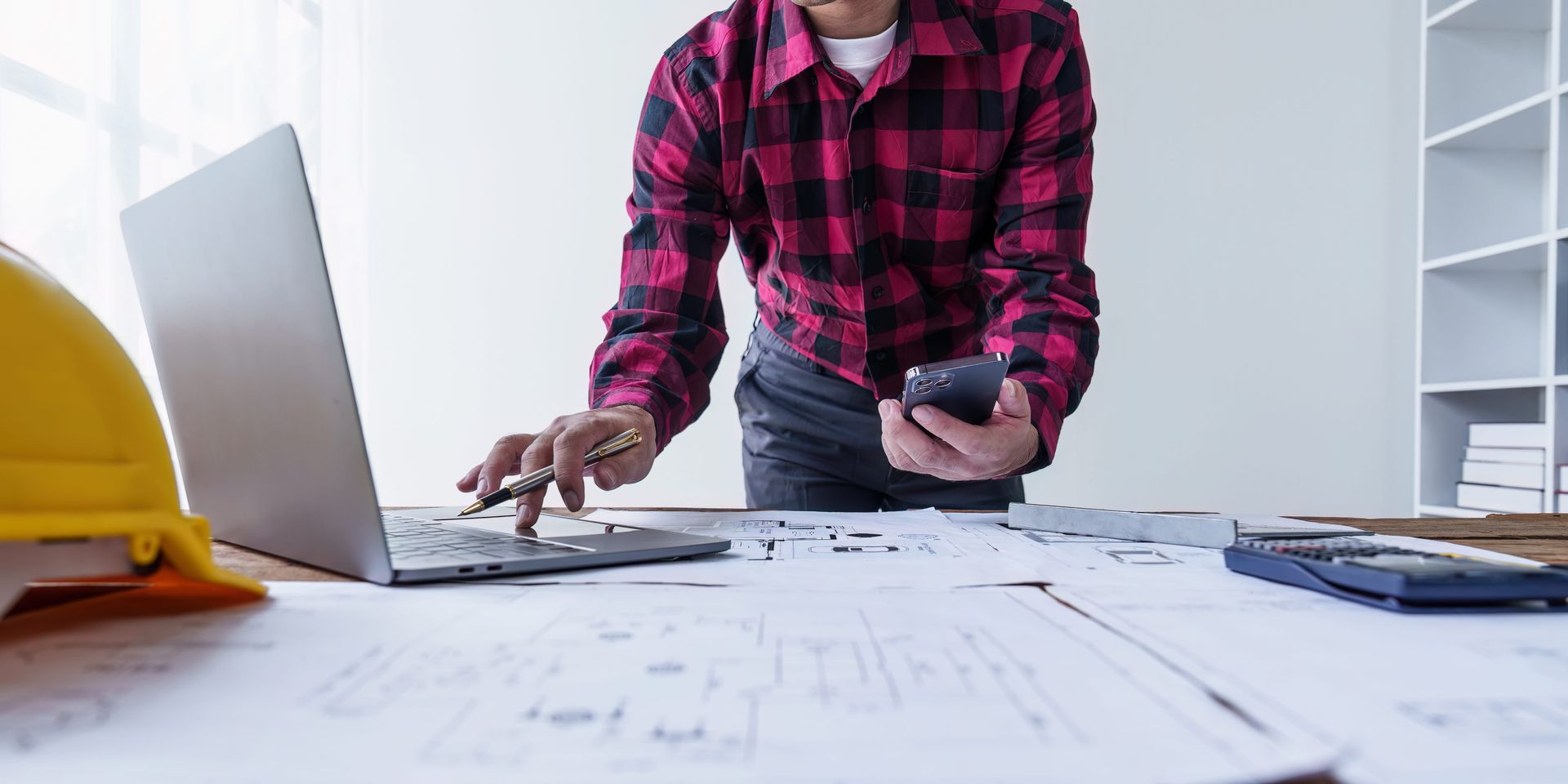 Person in a pink plaid shirt works at a desk with laptop, papers, and hard hat in a bright office.