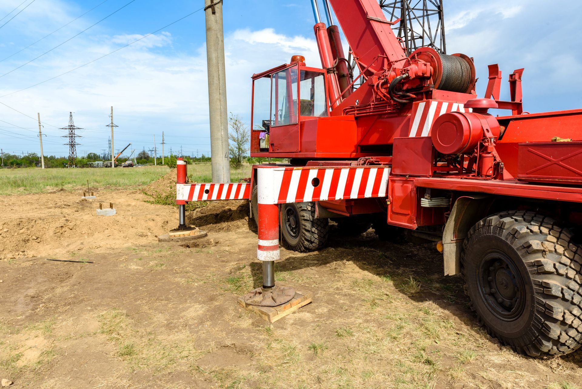 Red crane truck with striped outriggers parked on dirt near utility poles under a blue sky