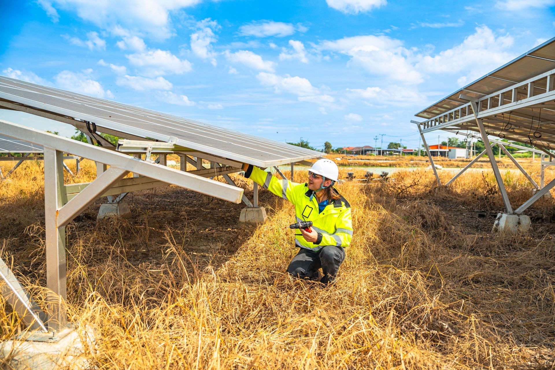 Worker in neon safety gear kneeling in dry grass beside a solar panel array under a blue sky