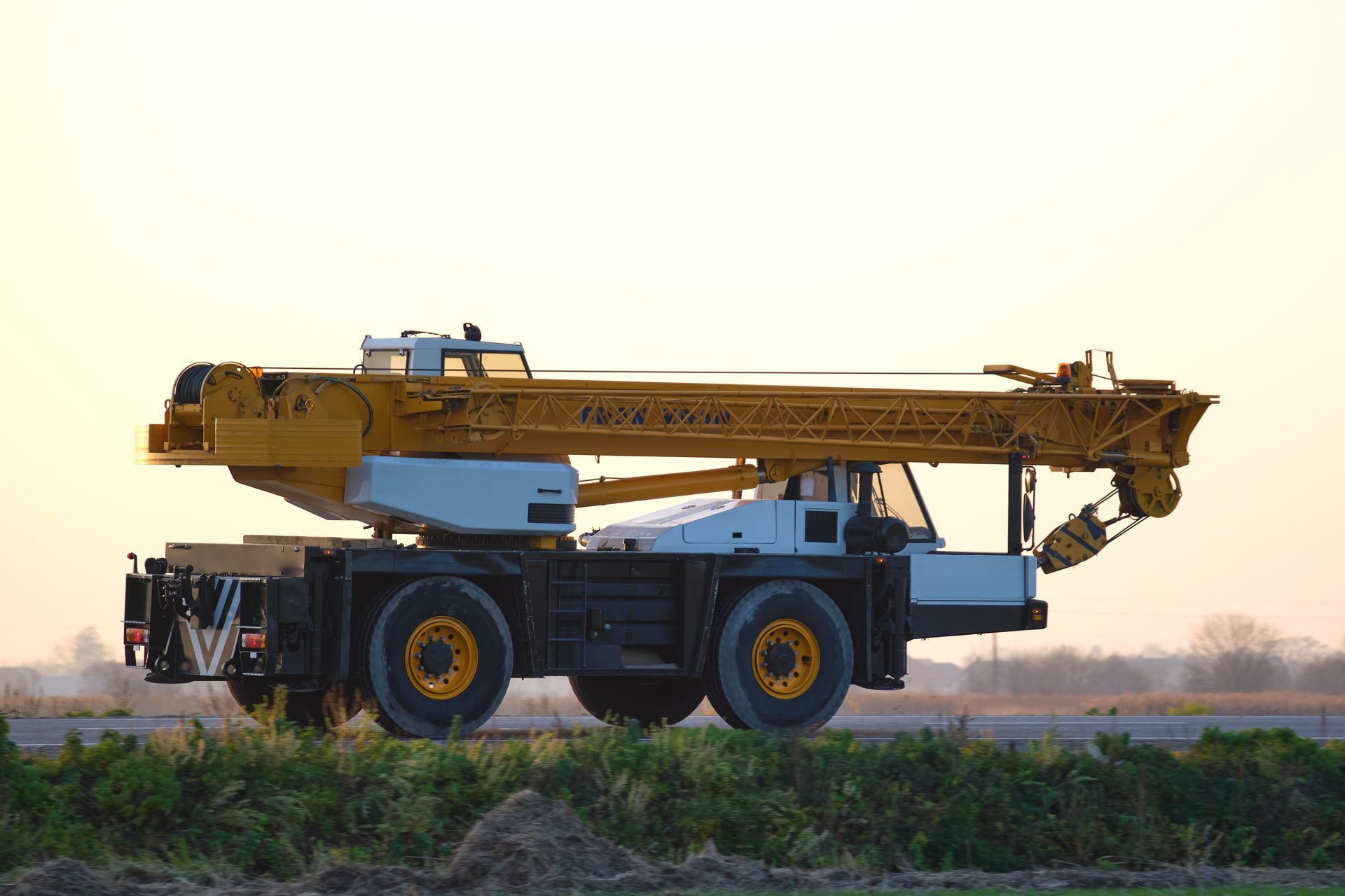 Yellow mobile crane truck on a dirt road at sunset, with long boom extended.