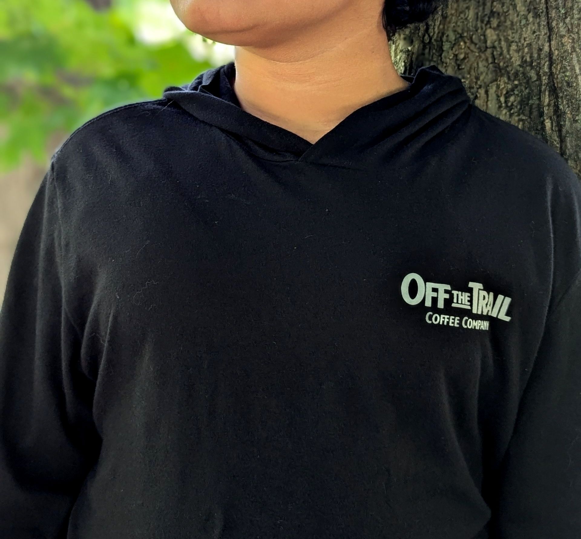 A person wearing a black off the trail hoodie leans against a tree