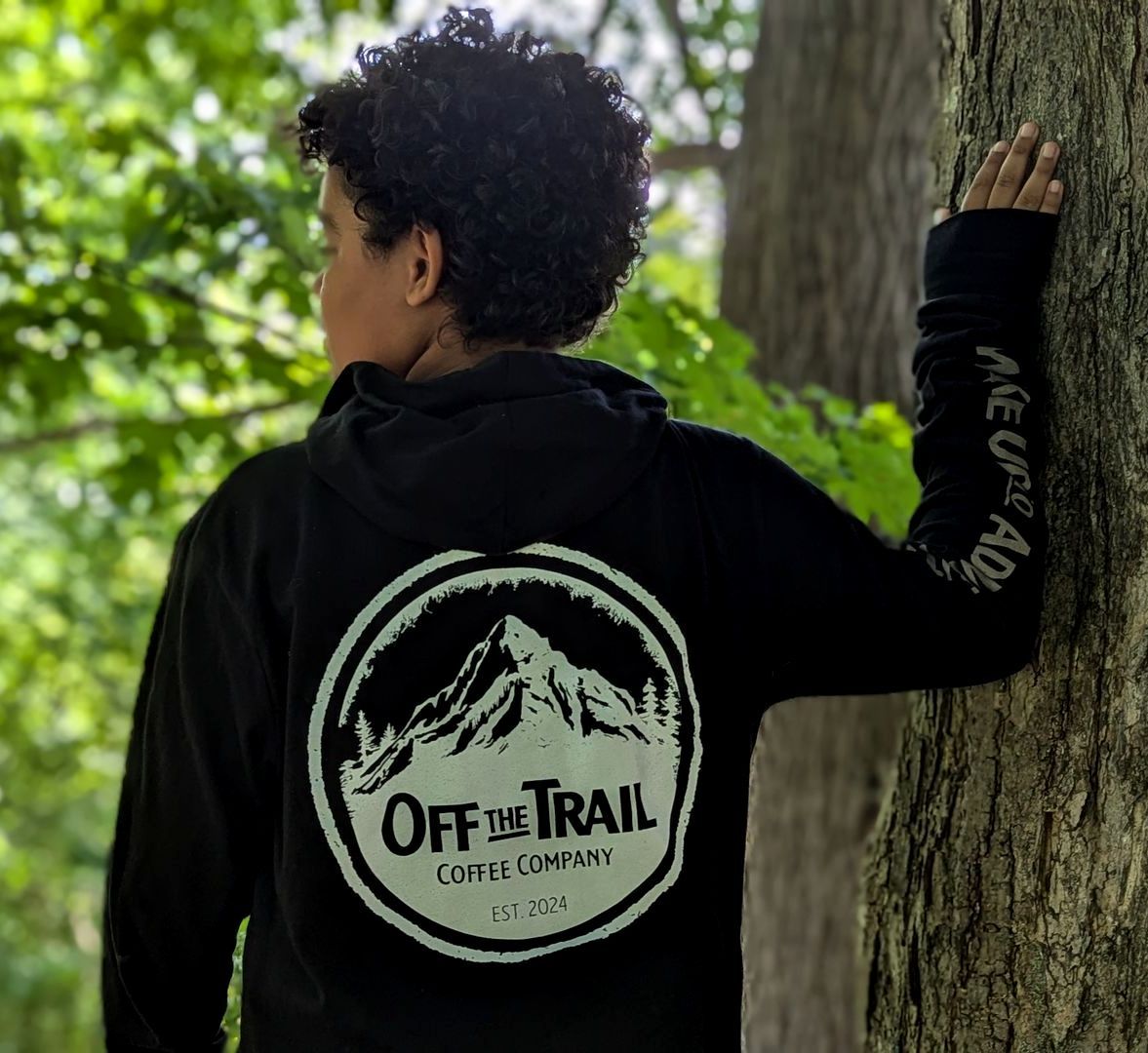A person wearing a black off the trail hoodie leans against a tree