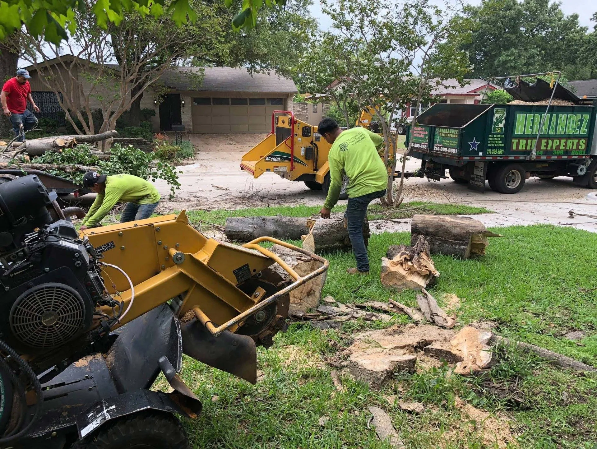 Workers clear storm debris with yellow machinery and a green truck on a muddy residential street.