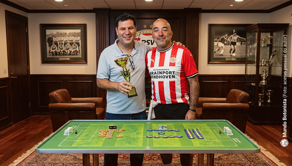 Dois homens estão de pé atrás de um jogo de futebol de mesa, um deles segurando um troféu, em uma sala decorada com objetos esportivos.