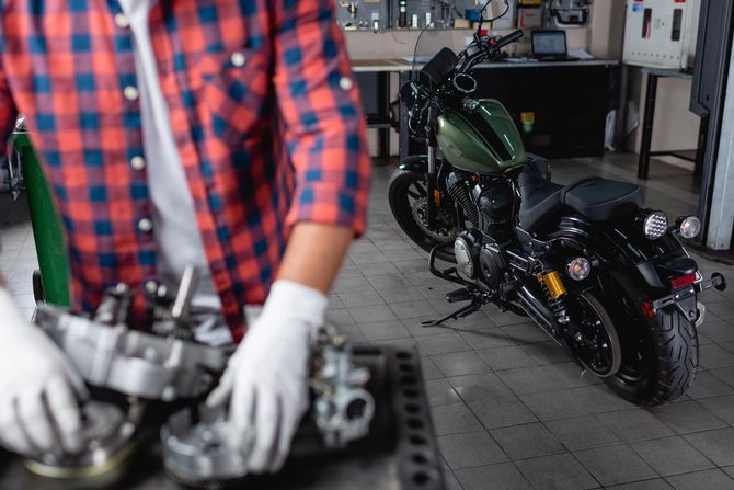 Meccanico con camicia a quadri rossi e guanti al lavoro su componenti di un motore in un'officina, con una motocicletta sullo sfondo.