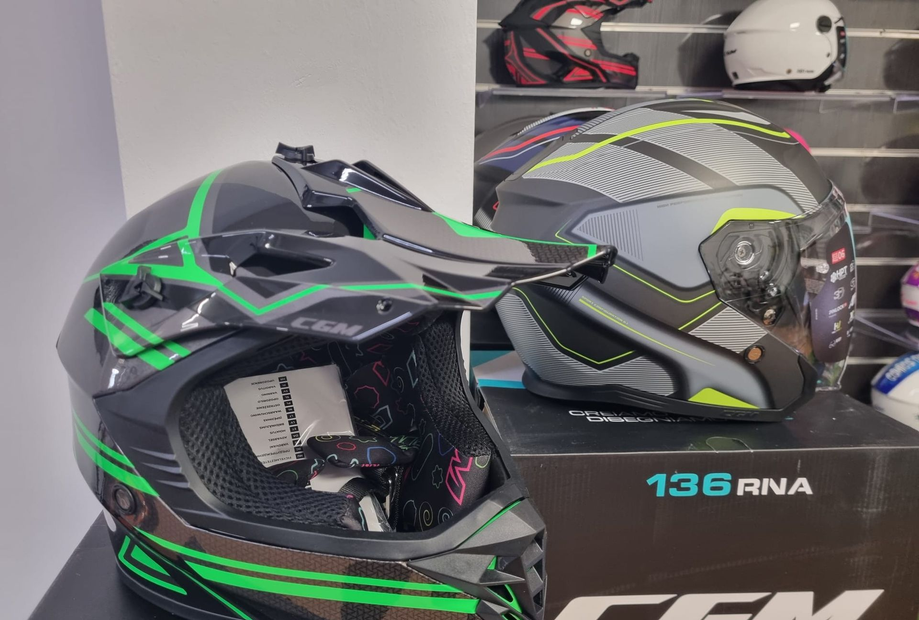 Un casco da motocross nero e verde accanto a un casco grigio su uno scaffale, con un casco bianco sullo sfondo.