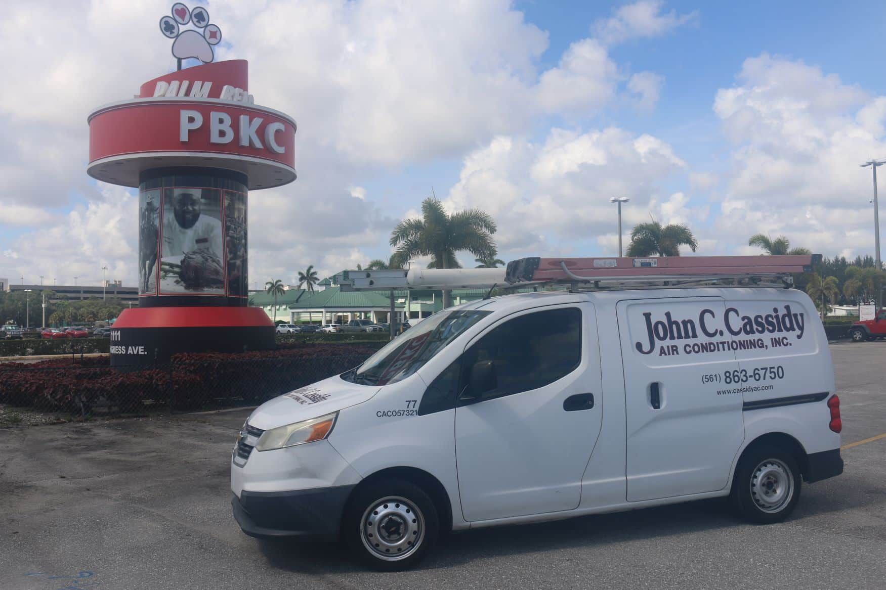 White John C. Cassidy A/C van parked in front of the Palm Beach Kennel Club sign. Cloudy sky in background.