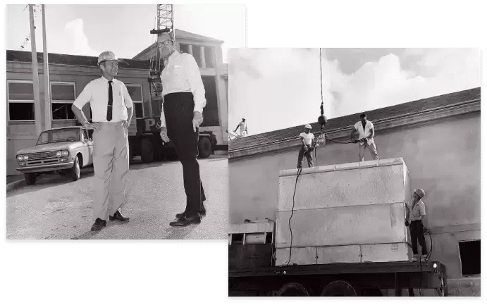 Two men watching the installation of a large object onto a building. A car is parked nearby.