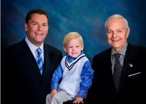 Three generations in suits: man, child, and older man, smiling in front of a blue background.
