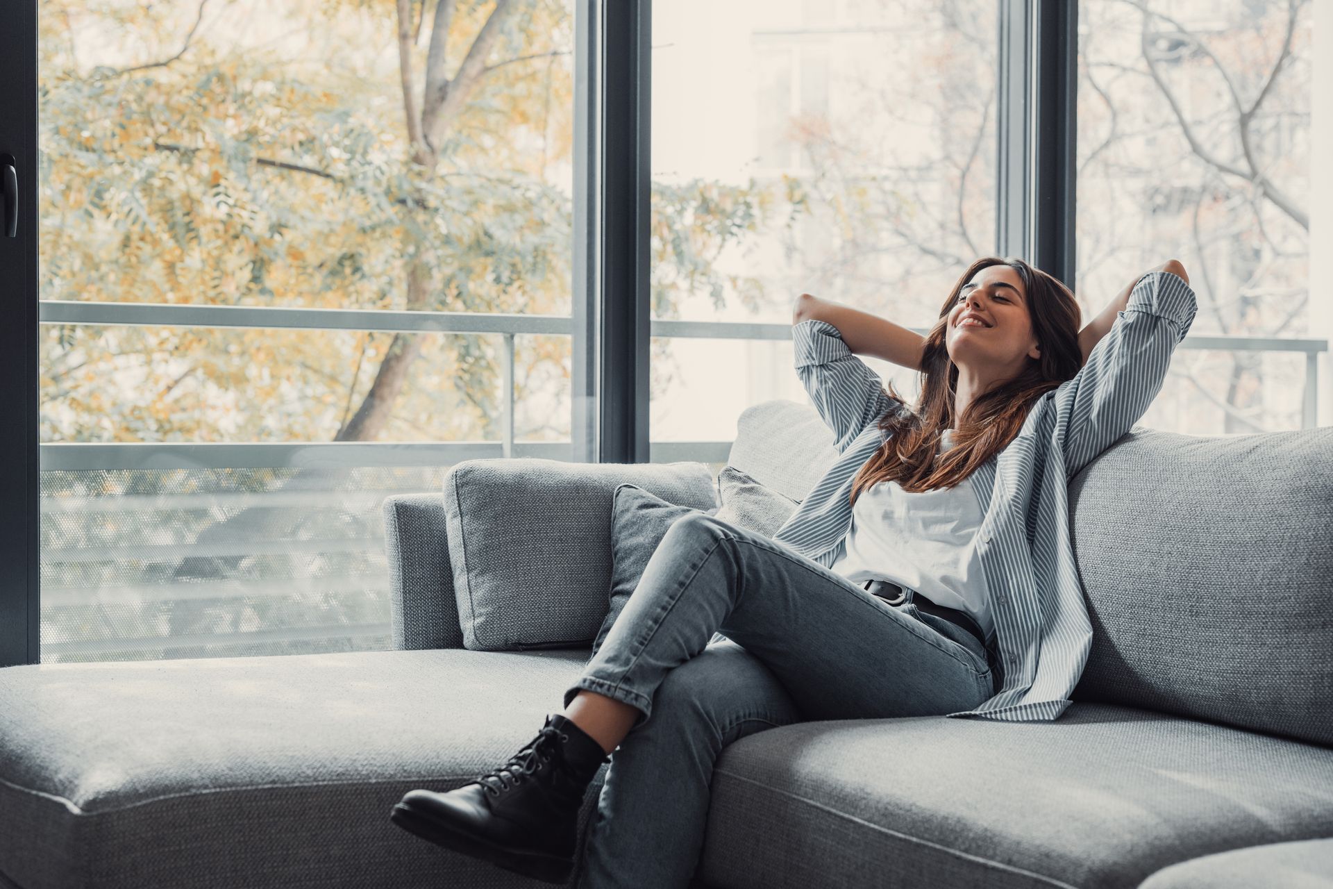 Woman relaxing on a grey sofa, hands behind head, smiling. Large window with outdoor view.