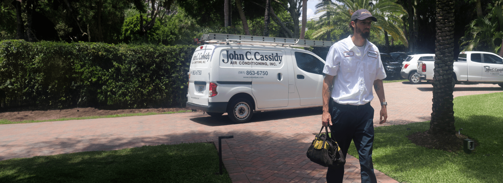 Man in uniform walking toward the camera, carrying a toolbox, by a white work van parked on a paved driveway.