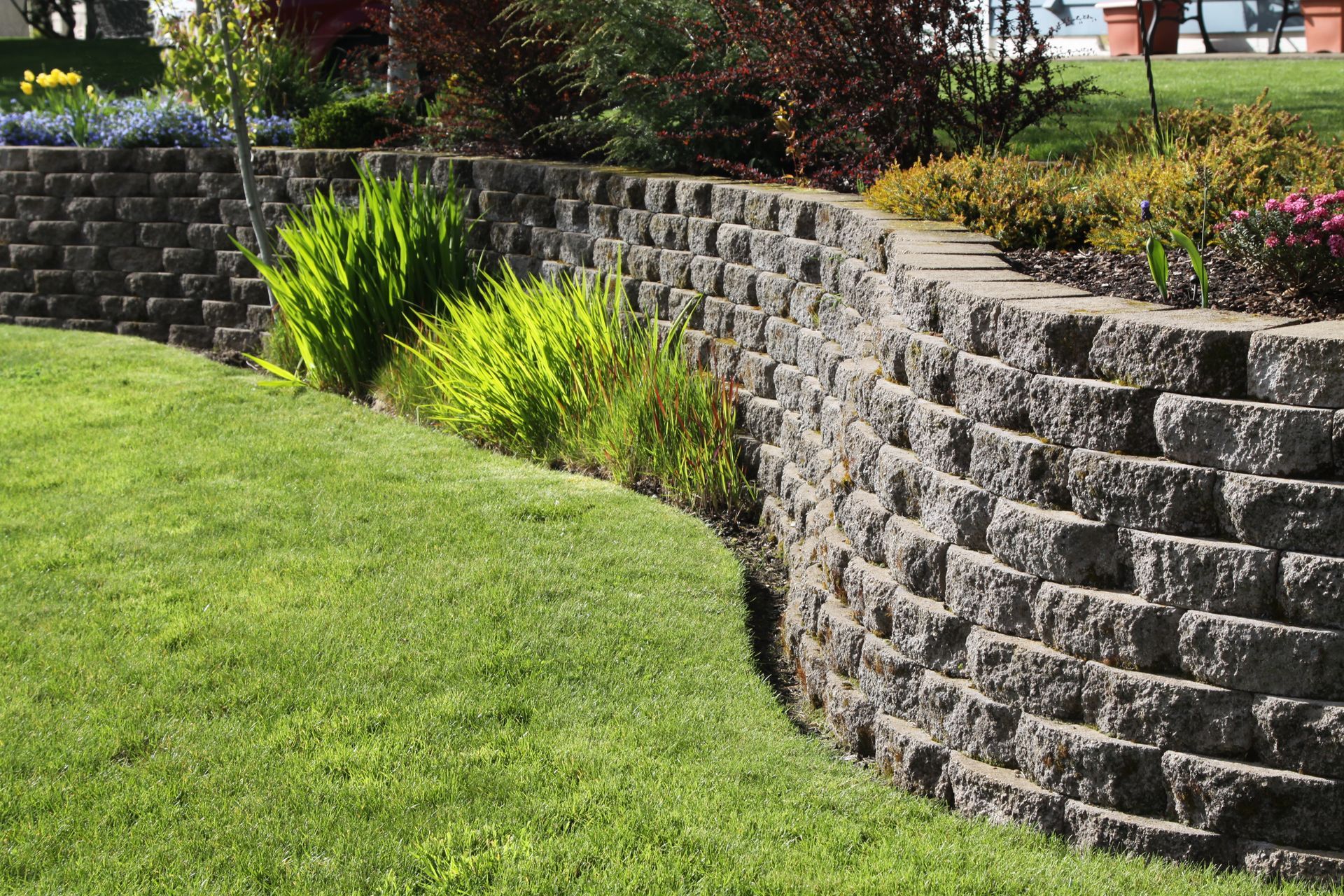 Curved retaining wall made of gray stone blocks, separating green lawn from garden with shrubs and flowers.
