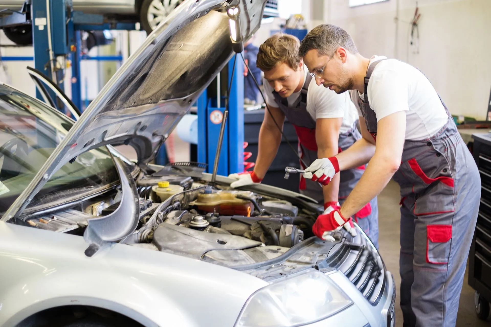 Two mechanics inspect a silver car engine in a repair shop, hood open