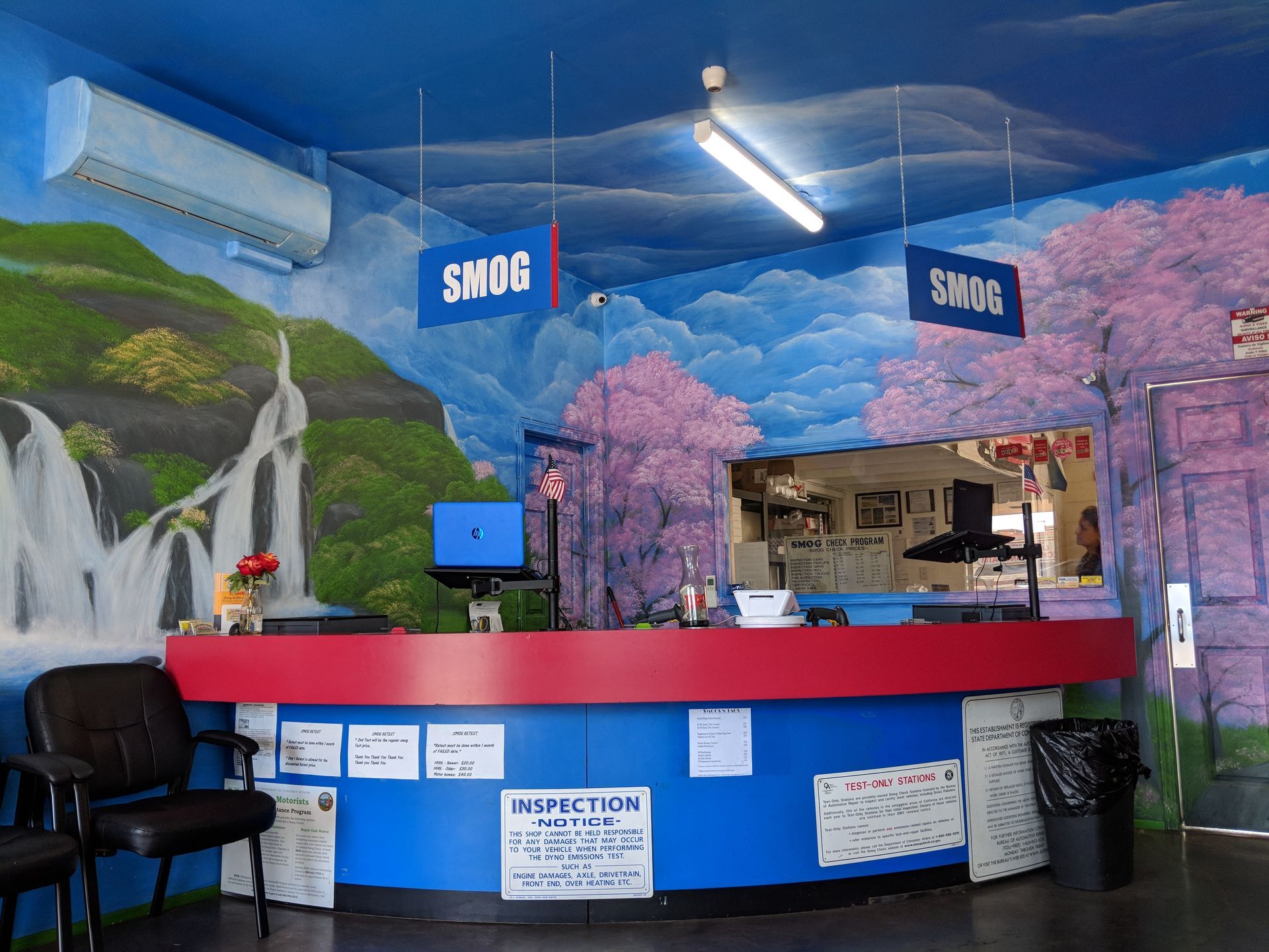 Empty reception desk with blue-and-red counter, cherry blossom murals, and hanging SMOG signs inside a colorful lobby