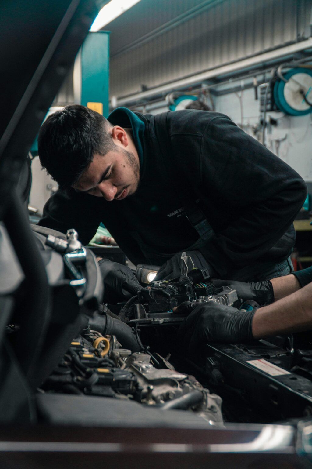 Mechanic inspecting engine under hood, focused in a dim garage with tools and blue overhead lights