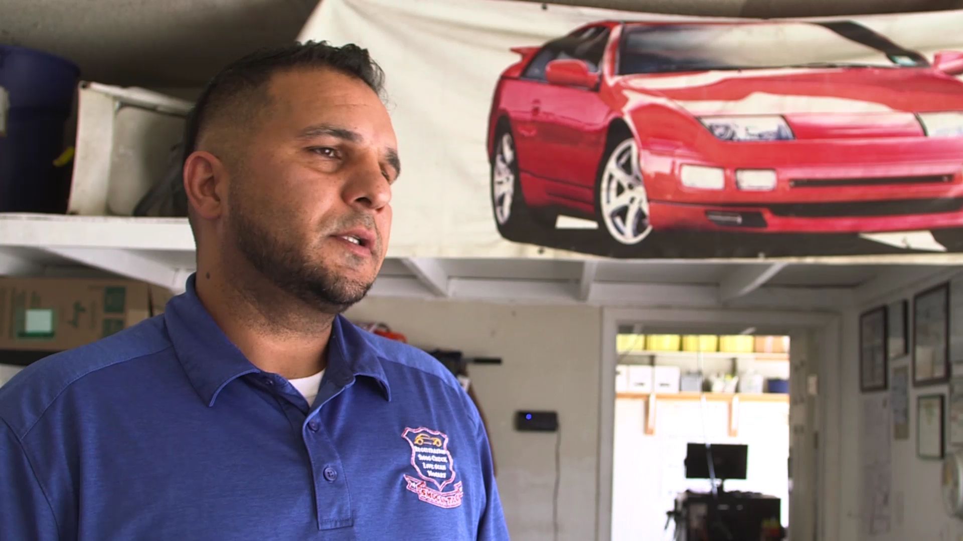 Man in blue shirt inside a garage, standing beneath a banner showing a red sports car