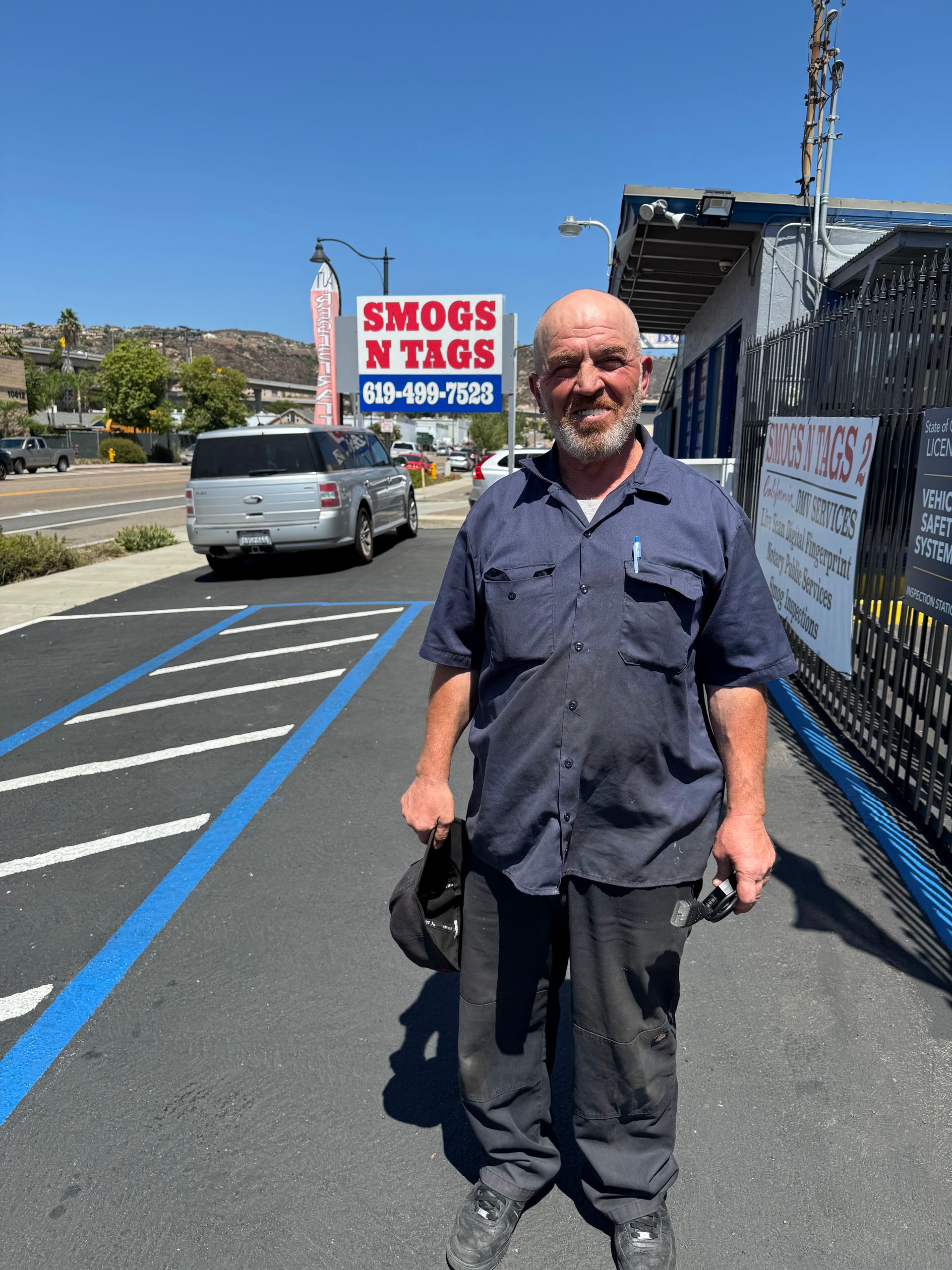 Man standing on a sunny sidewalk beside a truck and fence with a “Shog’s Towing” sign behind him.