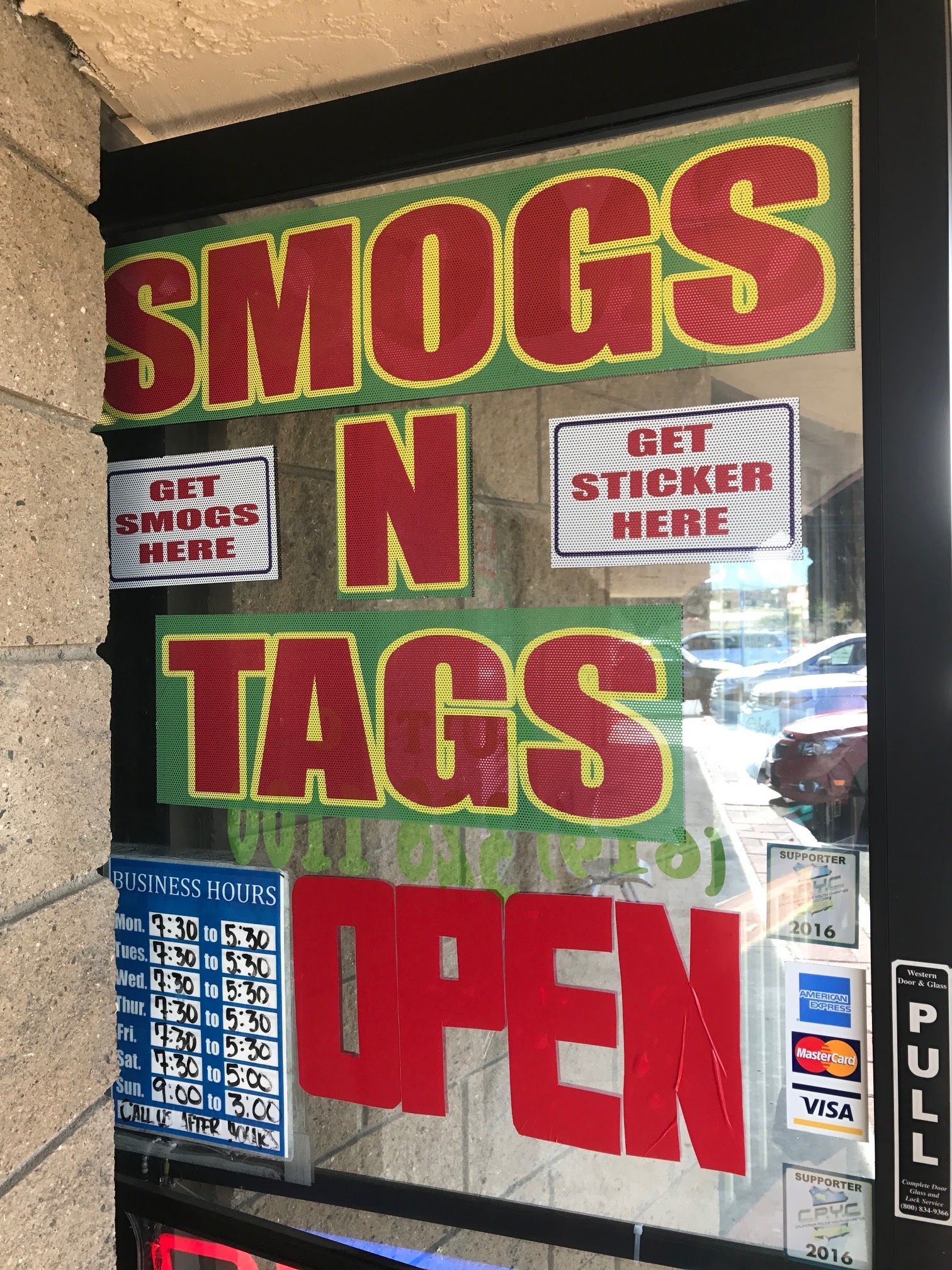 Storefront window sign reading “SMOGS N TAGS OPEN” in red and green, with “Get stickers here” notices.