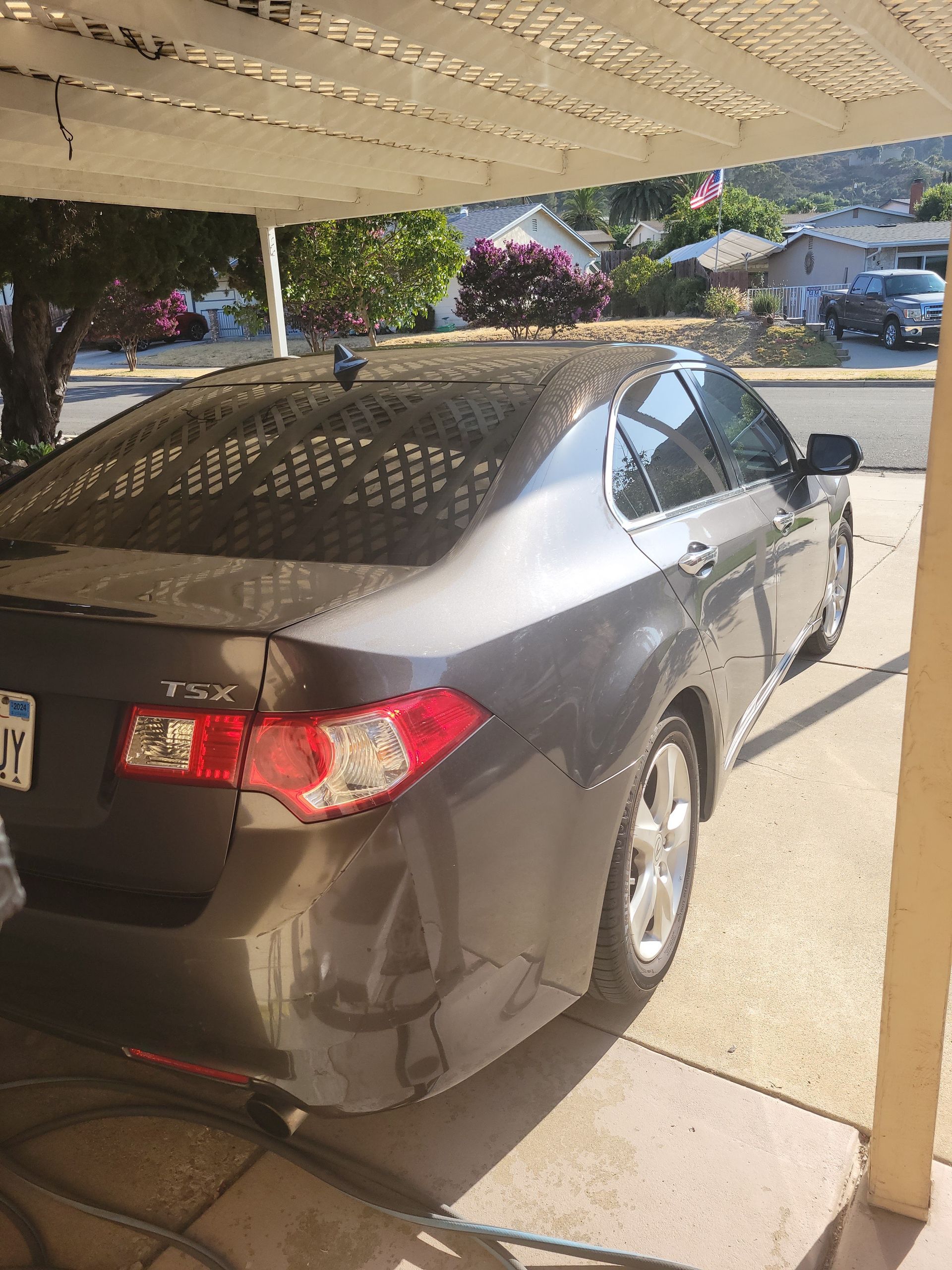 Dark gray sedan parked under a canopy in a driveway, rear and side view, with sunlight and tree shadows.
